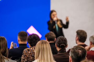 Obraz premium Speaker presents to an engaged audience at a conference. Bright blue backdrop and geometric light add energy to the scene. Attendees sit and listen, some recording on devices.