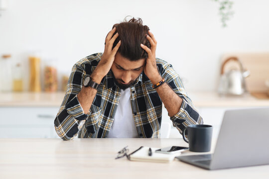 Depressed arab man sitting in front of laptop at home, touching his head, suffering from burnout or financial crisis, young middle-eastern freelancer having problems at job, copy space