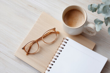 A pair of glasses and a coffee mug sit on a wooden table next to a notebook