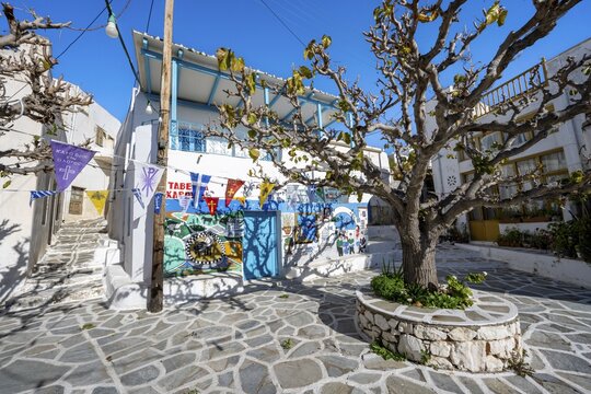 Small square with painted houses, trees and colourful flags, alleys of the village Marpissa, Paros, Cyclades, Greece