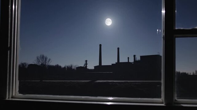 Industrial landscape viewed through a window with the full moon shining in the clear night sky. The factory silhouette stands against the dark blue sky