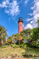 A red lighthouse stands tall in front of a lush green forest. The sky is clear and blue, and the trees are full and green. The scene is peaceful and serene