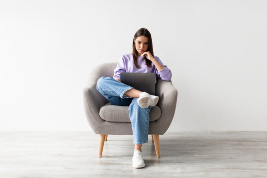 Serious young lady sitting in armchair with laptop computer, studying remotely or video conferencing against white studio wall, full length. Millennial woman watching webinar, having online job