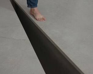 A bare foot wearing denim approaches the sharp edge of a linear, dark void separating gray tiled flooring.