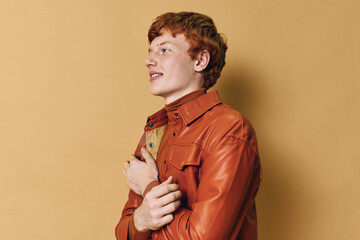 Redhead young man wearing orange leather jacket posing on beige background with confident expression, casual style and side profile portrait in studio light. © SHOTPRIME STUDIO