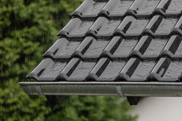 Roof corner with black tiles and rain gutter in heavy rainfall
