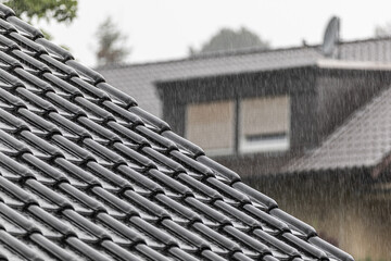 Diagonal roof edge with black tiles in heavy rain, blurred neighbor house
