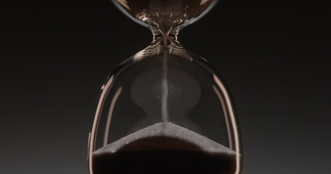 Closeup of hourglass measuring time over a dark dramatic background. Macro view of sand falling down inside the glass clock counting minutes. Concept of time and life flowing.