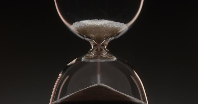 Closeup of hourglass measuring time over a dark dramatic background. Macro view of sand falling down inside the glass clock counting minutes. Concept of time and life flowing.