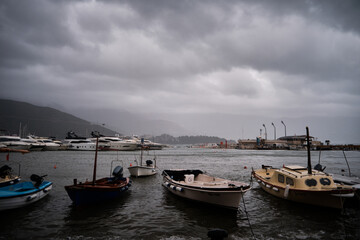 Small boats and fishing vessels float in coastal harbor under dark overcast skies. Choppy water and distant piers create moody maritime scene. Hills faintly visible in background. Budva, Montenegro