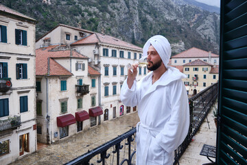 Man in white bathrobe and towel on his head stands on balcony overlooking historic European old town and drinks from wine glass. Buildings with shuttered windows and red roofs at background