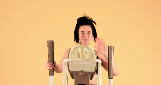 A young woman exercises on an elliptical trainer and waves hello with a smile on her face in this high-quality 4K video. The footage conveys positivity, fitness, and an active lifestyle, making it ide
