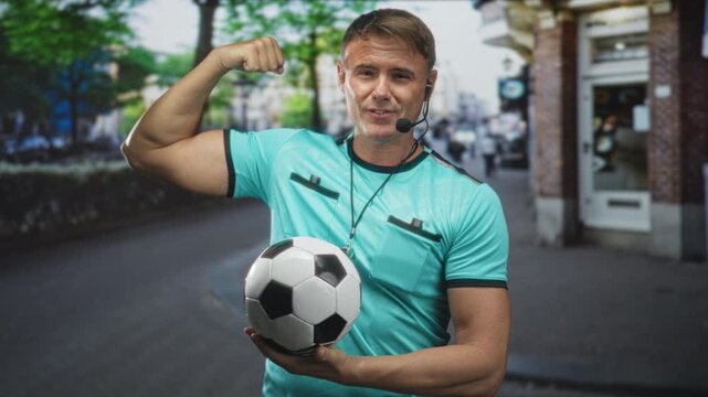 Man referee in turquoise shirt holds soccer ball, wears whistle and headset, flexes arm and smiles on street; confidence.