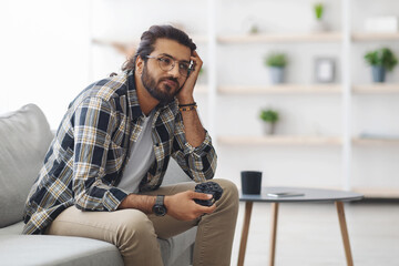 Bored indian guy sitting on couch with joystick, looking at copy space, playing video games alone at home. Middle-eastern young man feeling lonely while spending weekend alone, home entertainment