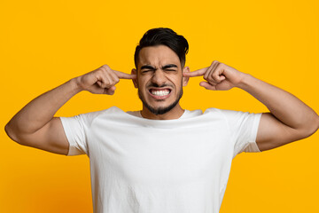 Annoyed millennial middle eastern guy in white t-shirt covering his ears over yellow studio...
