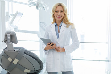Female dentist pretty blonde woman keeping beautiful smile on her face and holding digital tablet...