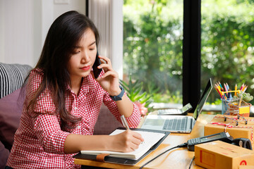 Young Asian entrepreneur talking on mobile phone and writing customer orders. SME online business owner working at home with laptop and parcel boxes.