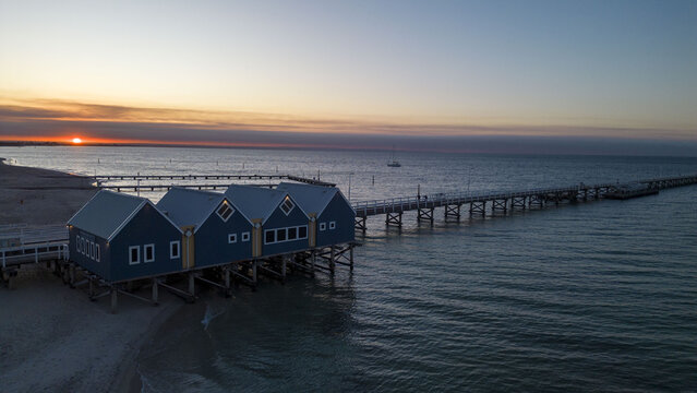 Aerial view of the iconic blue Busselton Jetty bathed in the warm glow of the setting sun against the tranquil, darkening ocean, Busselton, Western Australia, Australia.