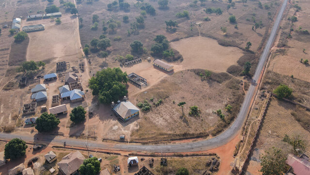 Aerial view of muted ochre earth meeting the dark asphalt road curving through sparse trees, a stark contrast to the clustered buildings., Essan, Niger, Nigeria.
