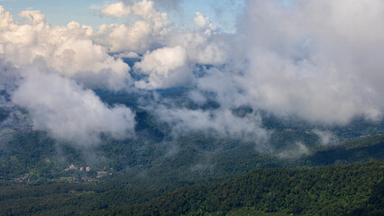 Aerial view of green mountain valley covered with white clouds and mist. Small village located in deep forest nature landscape.