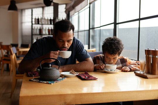 African American man and his son are having lunch in cafe at table, teenager and his father are sitting in restaurant and eating and drinking coffee