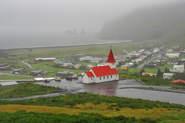 Rainy and cloudy day in Iceland. Blurry view. Wet landscape. Panoramic view to Vik small Icelandic seafront village. V&iacute;k &iacute; M&yacute;rdal Lutheran church on the hill, is white building with red roof and tower