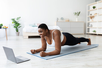 Online sports and weight loss. Overweight young black woman doing exercises in front of laptop at home, full length. Plus size African American lady standing in elbow plank, following web tutorial