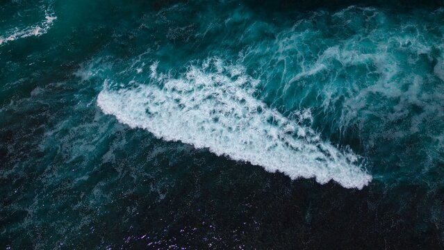 Aerial view of the dark turquoise sea crashing into white foam waves, creating a mesmerizing contrast of colors and textures, Bali, Bali, Indonesia.