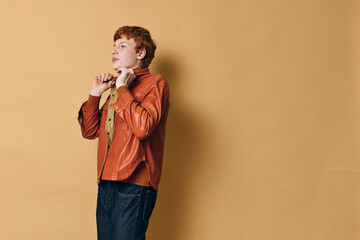 Young man with red hair adjusting collar of brown leather jacket standing against beige background. Casual style, confident posture with thoughtful expression and modern outfit for fashion concept.