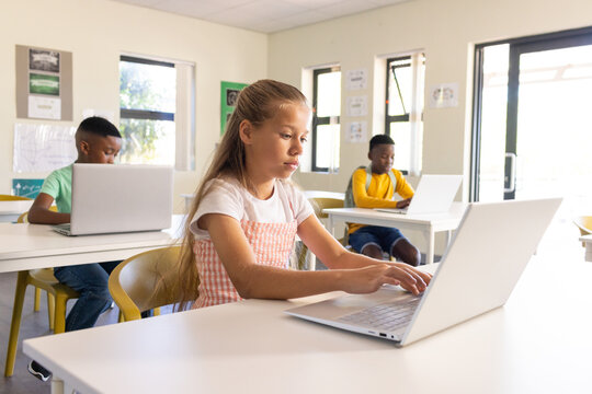 Diverse children at school typing on silver laptops at white desks, girl wearing pink gingham dress