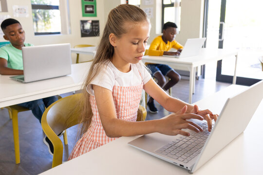 Classmates typing at white desks using silver laptops in classroom with girl in pink gingham dress