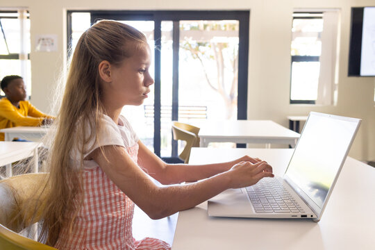 Diverse classmates with female child in pink gingham dress using silver laptop in classroom