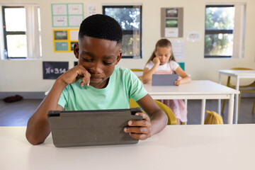 African American classmates sitting at white desks in primary classroom, holding dark gray tablets