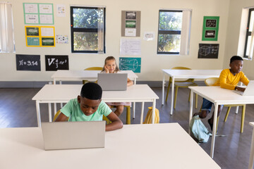 Diverse youth students sitting at white desks in classroom, using laptops and viewing math posters
