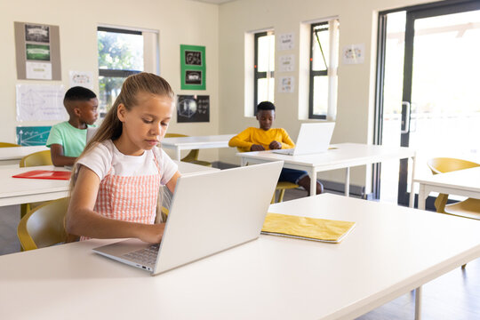 Diverse classmates sitting at white tables using silver laptops, female youth with yellow notebook