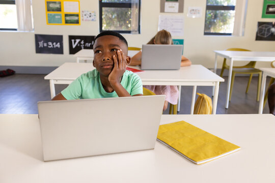 African teen student sitting in class at desk using silver laptop with yellow notebook looking up
