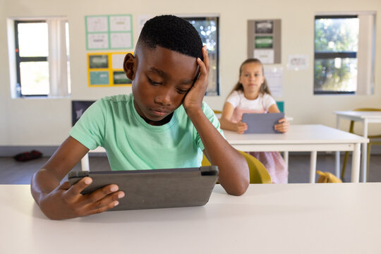 Diverse children sitting in class at tables with African American boy in mint tee using tablet