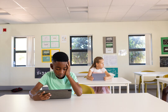 Students sitting at white desks using tablets in classroom with posters, yellow chairs, windows