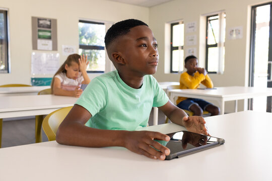 Diverse youth classmates learning at white desks under windows, boy in light green tee using tablet