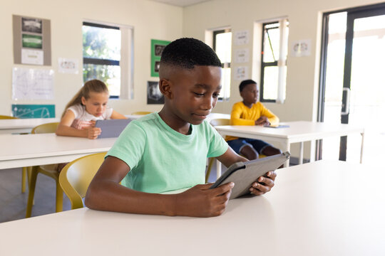 Diverse children sitting in classroom, holding tablets and working at white desks near windows