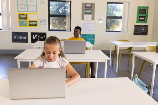 Diverse child classmates using silver laptops at school desks with math posters showing ?=3