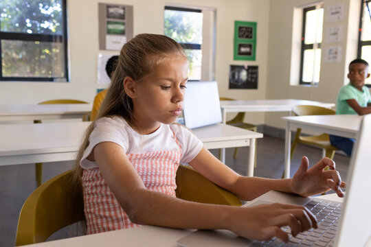 Diverse classmates including female 10-12yr child wearing pink pinafore using laptop in classroom