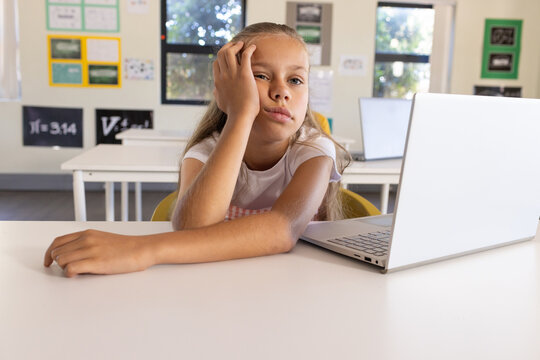 Teenage girl sitting at classroom table, resting head on hand, using silver laptop