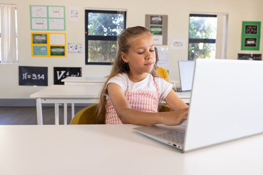 Diverse classmates, female child in pink pinafore working in classroom at desk using silver laptop