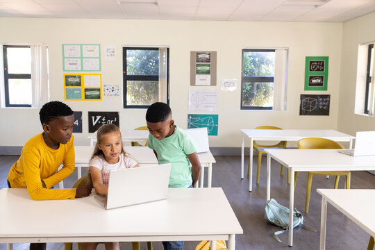 Diverse classmates leaning around silver laptop in bright classroom, sharing schoolwork and notes
