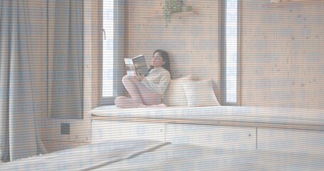 Sitting woman wearing knit sweater, pants on built-in daybed drawers at bedroom nook, reading book