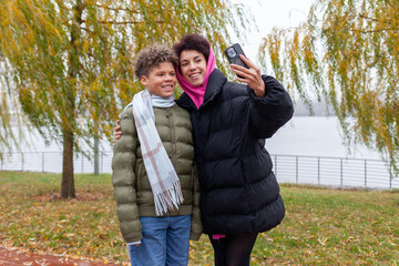 African American woman and her son take selfie in park in autumn, mother and child in warm jackets take photos on smartphone outdoors
