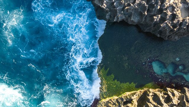 Aerial view of the dramatic clash of turquoise waves against the rugged, dark cliffs edges, Nusa Penida, Bali, Indonesia.