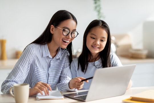 Happy asian mom helping her studying daughter, looking at laptop screen together, sitting in kitchen interior. Excited school girl using computer, having online lesson while coronavirus pandemic