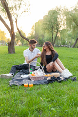 young couple in love is sitting on blanket in the park at picnic and eating, girl and guy are drinking juice and relaxing in nature on date